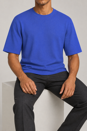 Seated studio view of Open Hours Knit T-Shirt in Royal, showing blue knit drape above charcoal trousers.