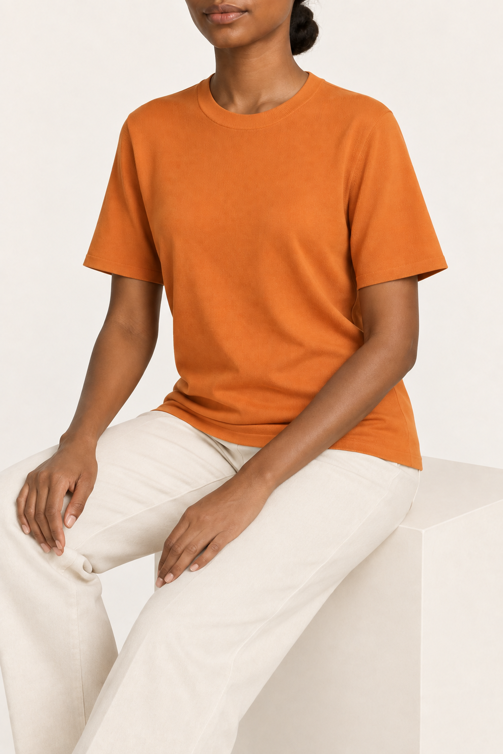 Seated studio view of Soft Tempo Knit T-Shirt in Orange, showing orange knit drape above cream trousers.