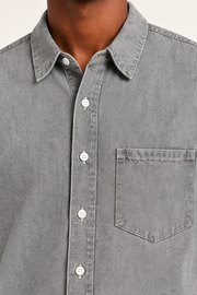 Detail of Worn Line Denim Shirt in Light Gray, showing the pointed collar, white front buttons and stitched patch pocket.