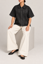 Seated view of Soft Shift Zip Denim Shirt in Black, showing the loose silhouette and natural drape over ivory trousers.