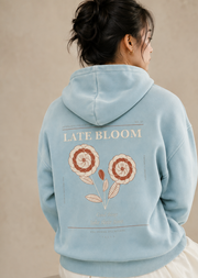 Seated rear three-quarter studio view of the TELLS Late Bloom Poster Hoodie in Lake Blue with botanical back print.