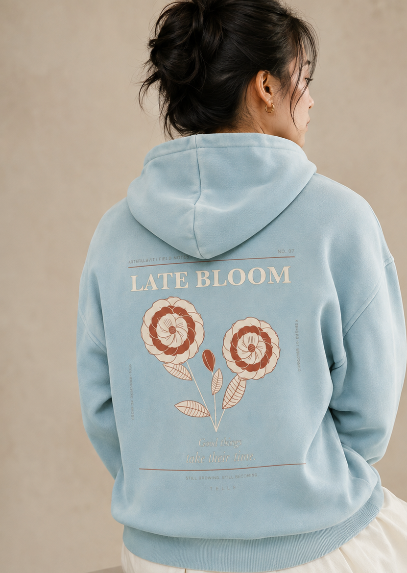 Seated rear three-quarter studio view of the TELLS Late Bloom Poster Hoodie in Lake Blue with botanical back print.